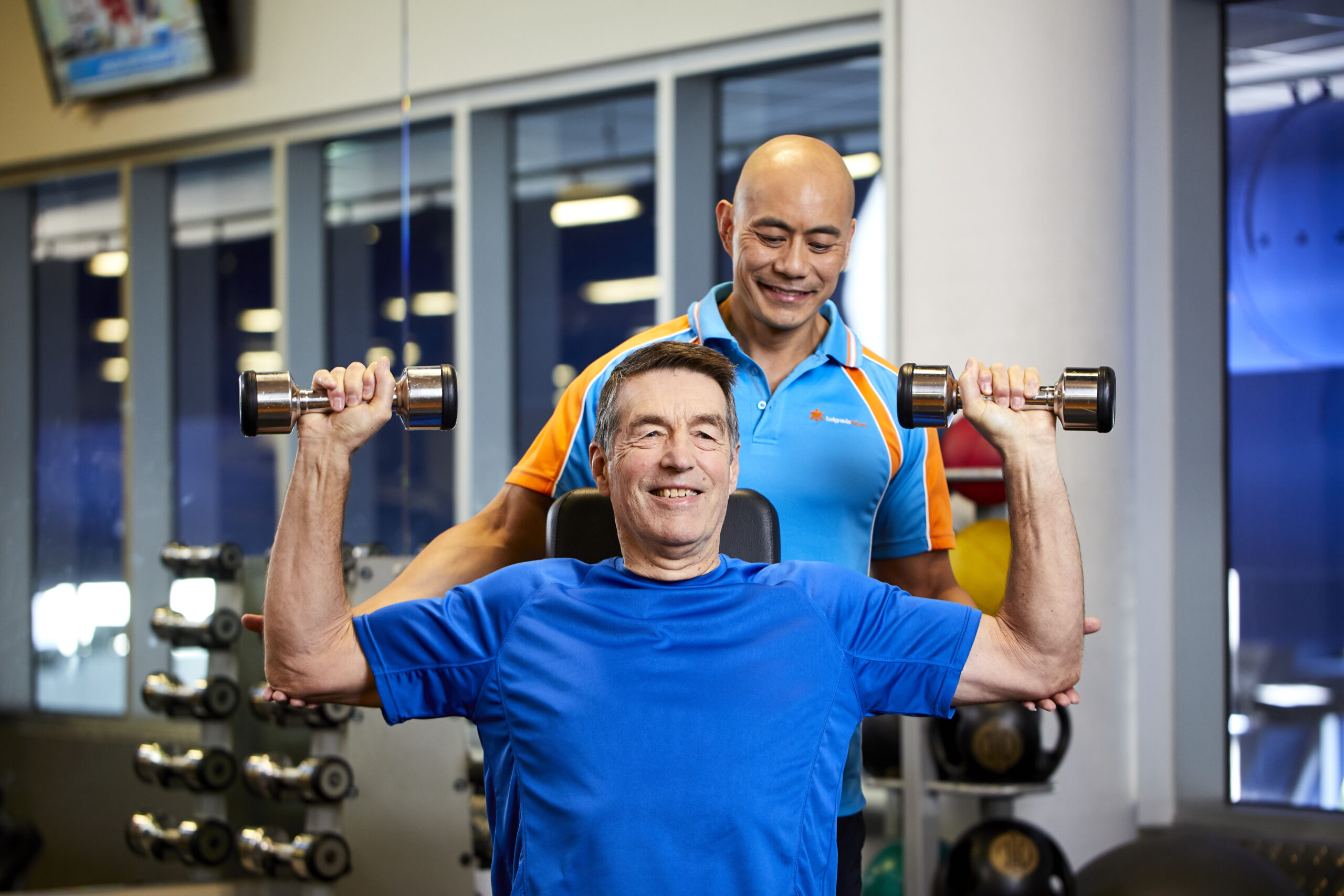 A gym trainer assists an older man with lifting 2 dumbbells.
