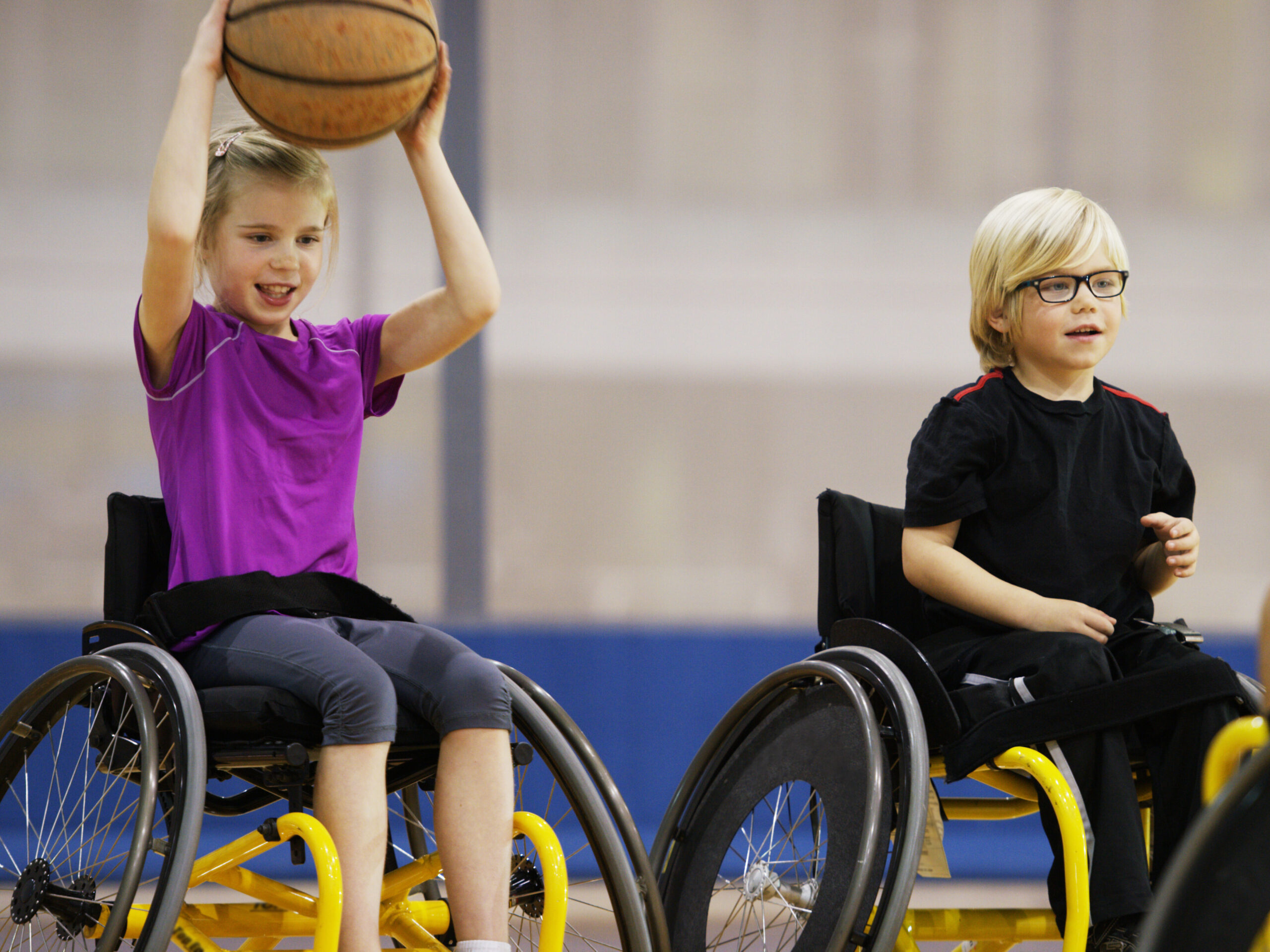A little girl in a wheelchair is tossing a basketball to her athletic coach in the gym.
